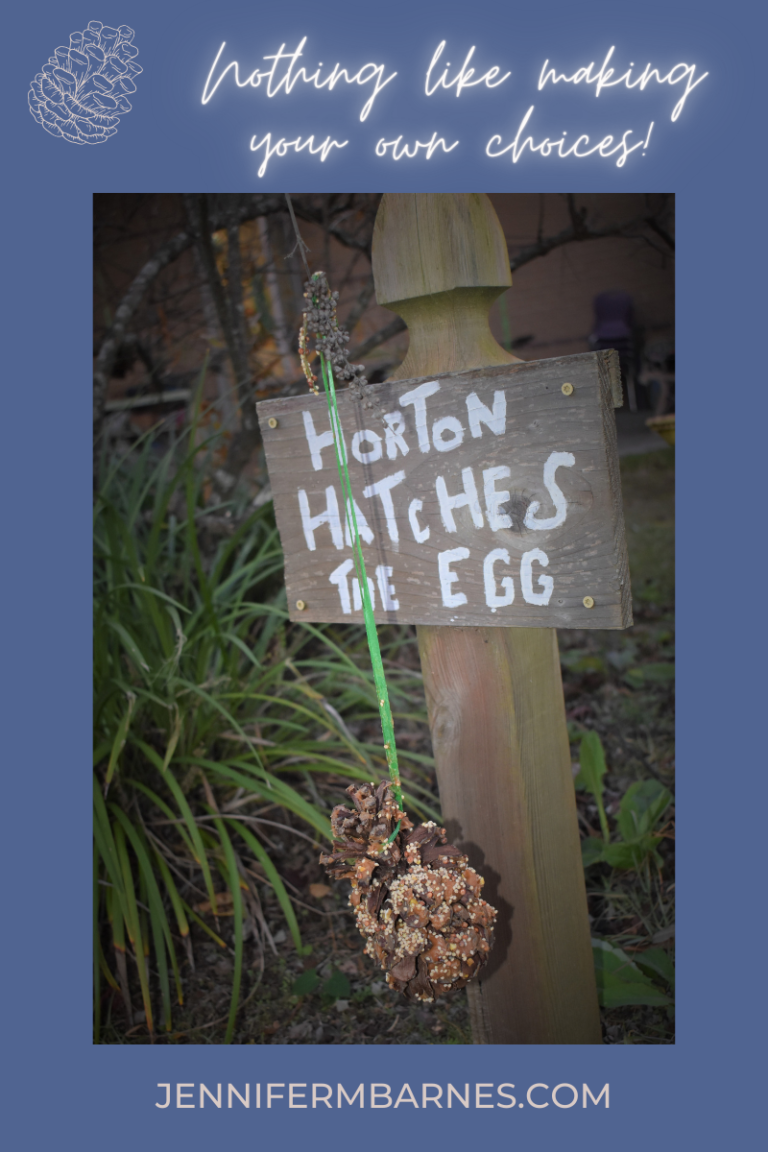 Close-up of a garden sign with a peanut butter pinecone decoration hanging on it. Words above state, "Nothing like making your own choices!"