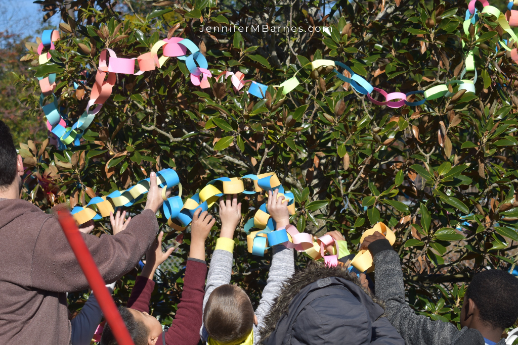 Image of children reaching to place a long paper chain around their own Night Tree with the help of an adult.